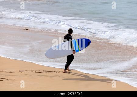 Surfeur transportant une planche de surf, Playa Piedra surf Beach, El Cotillo, Fuerteventura, Îles Canaries, Espagne. Banque D'Images