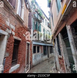Une calle (rue) étroite et calme à Venise, Italie, à la fin octobre 2024. Des bâtiments vénitiens traditionnels avec des fleurs et des volets fermés bordent le mouillé Banque D'Images