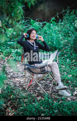 Heureusement jeune femme assise sur la chaise de camping portant casque écoutant de la musique de l'ordinateur portable tout en se relaxant sur le camping dans la forêt Banque D'Images