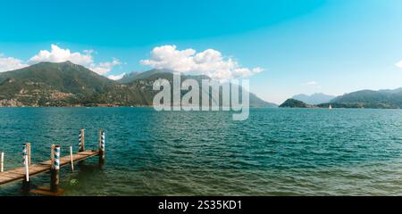 Scène tranquille d'une jetée en bois s'étendant dans les eaux calmes du lac de Côme, entouré de montagnes verdoyantes et d'un ciel bleu clair, créant une paix Banque D'Images
