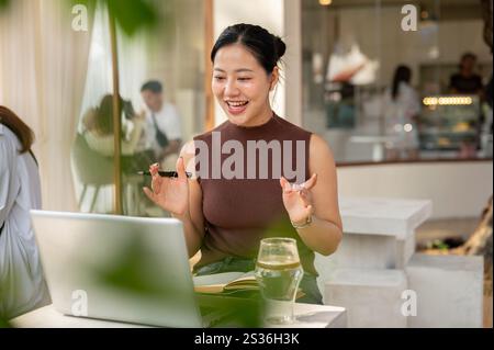 Une femme asiatique heureuse et confiante travaille à distance depuis un café et participe à une réunion en ligne avec ses collègues via son ordinateur portable. femme d'affaires, Banque D'Images