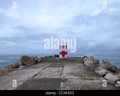Un phare rouge et blanc se dresse fièrement au sommet d'une jetée robuste, surplombant l'eau environnante. Phare rouge et blanc au sommet de la jetée Banque D'Images