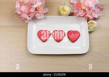 Trois biscuits roses et rouges en forme de coeur sur une assiette de service blanche sur une table en bois avec des fleurs de couleur rose et crème. copier l'espace Banque D'Images