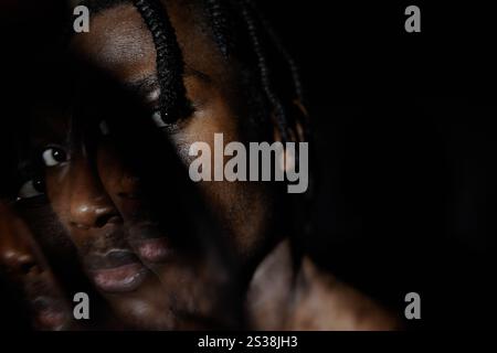 Portrait d'homme afro-américain regardant avec un regard perçant à la caméra cachant le visage dans l'ombre sombre profonde couvrant la moitié de son visage posant dans le studio sombre, effet de double exposition, espace de copie Banque D'Images