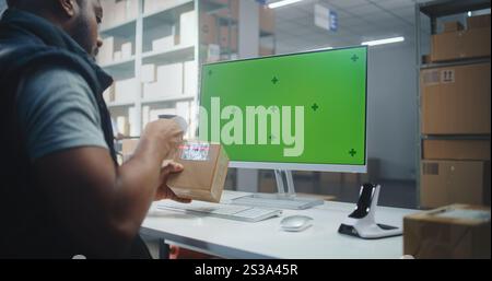 Service de livraison : African American Warehouse Associate scanne le code sur la boîte en carton avec scanner, entre les informations sur le colis sur ordinateur. L'écran de l'ordinateur affiche la maquette de la touche Chroma de l'écran vert. Portable. Banque D'Images