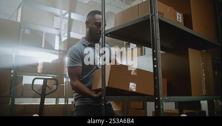 Le travailleur du centre de tri afro-américain décharge le transpalette, place les boîtes en carton, les colis sur les étagères. Entrepôt Retail Facility de Service de livraison avec E-commerce commandes en ligne, marchandises. Dolly Shot. Banque D'Images