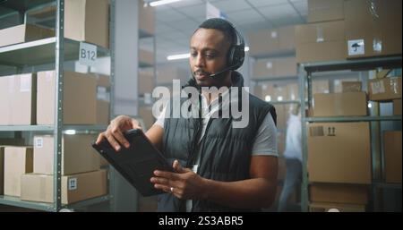 Afro-américain spécialiste de l'assistance à la clientèle en logistique dans le casque parle à l'appel avec le client, utilise un ordinateur tablette. Employé de l'entrepôt préparant les boîtes pour la livraison. Centre de tri des magasins de commerce électronique. Banque D'Images