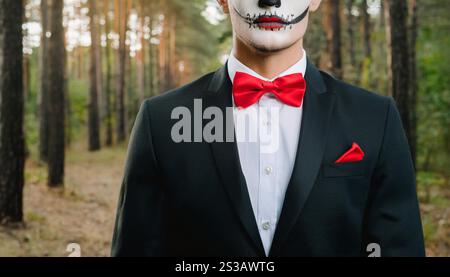 Un jeune homme en costume noir et noeud papillon rouge se tient dans une forêt, souriant avec un large sourire dentelé et portant un maquillage dramatique d'Halloween qui comprend Banque D'Images