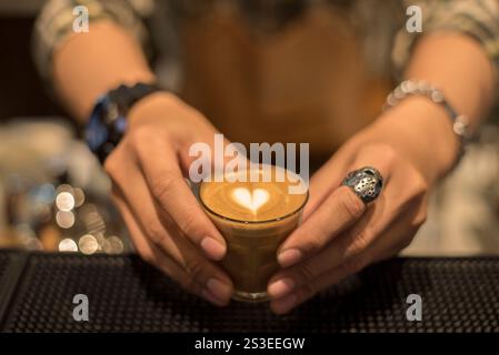 Barista tenant un petit verre de café avec latte art en forme de cœur, mettant en valeur le savoir-faire artisanal et l'ambiance chaleureuse du café. Banque D'Images