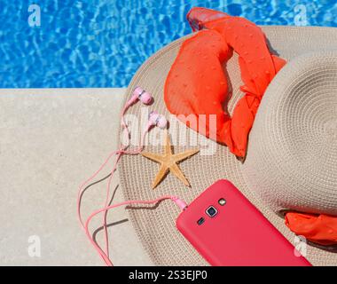 Chapeau de raphia pour femmes, téléphone et étoile de mer à côté de la piscine sur fond d'eau bleue. Banque D'Images