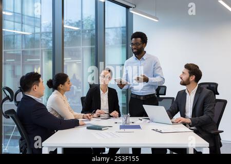 Réunion d'affaires, cinq collègues à l'intérieur du bureau dans la salle de conférence communiquent, un homme rend compte des résultats actuels des réalisations, de la détente dans une entreprise diverse personnes. Banque D'Images