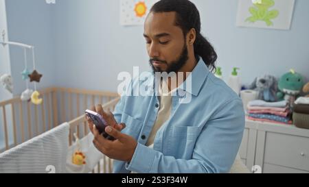 Homme dans la chambre tenant un smartphone près du berceau de bébé, entouré de jouets en peluche et de vêtements pliés, impliquant un environnement domestique confortable et attentionné. Banque D'Images