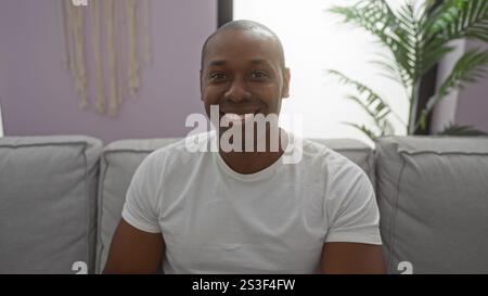 Jeune homme assis dans un salon élégant souriant portant une chemise blanche décontractée avec des plantes et un décor minimal, y compris l'art mural créant invitant et coz Banque D'Images