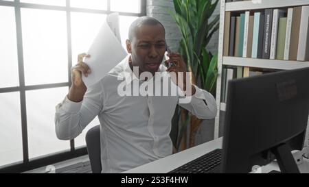 Homme afro-américain au bureau tenant des papiers et parlant au téléphone, paraissant stressé, avec des étagères et de grandes fenêtres améliorant le travail moderne Banque D'Images