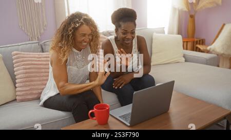 Des femmes assises ensemble dans un salon, riant et agitant devant un ordinateur portable, suggérant une réunion virtuelle ou un appel vidéo depuis la maison Banque D'Images