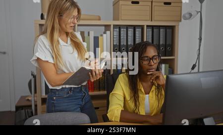 Jeune femme prenant des notes pendant qu'une autre femme travaille sur un ordinateur dans un cadre de bureau, mettant l'accent sur un environnement de travail collaboratif avec du mobilier de bureau Banque D'Images