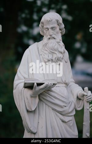 Gros plan extérieur d'une statue de pierre blanche représentant un homme barbu en robe, tenant un livre et un bâton. L'arrière-plan est flou, montrant foli vert Banque D'Images