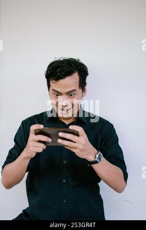 Un homme tenant un smartphone avec une expression excitée et concentrée, regardant attentivement l'écran. Parfait pour les thèmes comme le jeu, la technologie ou l'inte Banque D'Images