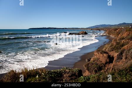 Les vagues s'écrasent contre un rivage rocheux à Cambria, en Californie, avec en toile de fond un ciel bleu clair et des collines lointaines. Banque D'Images