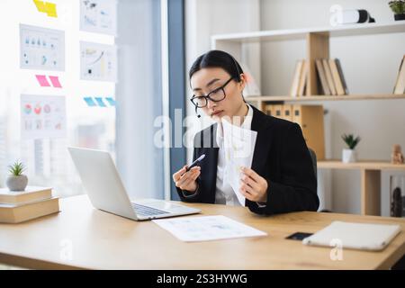 Femme d'affaires asiatique portant casque s'engage dans un appel vidéo en ligne tout en examinant des documents au bureau moderne. Banque D'Images