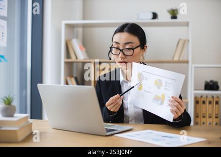 Femme d'affaires asiatique portant casque s'engage dans un appel vidéo en ligne tout en examinant des documents au bureau moderne. Banque D'Images