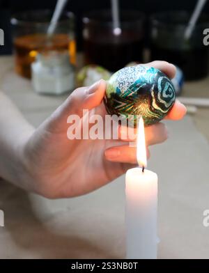 Photo de fabrication d'œufs de Pâques. Procédé pour enlever la cire d'oeufs peints. La main de la femme tient un oeuf au-dessus de la flamme de la bougie. La cire est partiellement éliminée du Banque D'Images