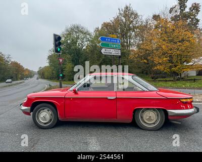 Une voiture rouge classique traverse une intersection tranquille entourée de feuillage d'automne dans une zone rurale Banque D'Images