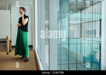 Une femme architecte mature converse sur son téléphone tout en se tenant debout dans un bureau contemporain avec de grandes fenêtres. Sa tenue vestimentaire et son comportement reflètent la profession Banque D'Images