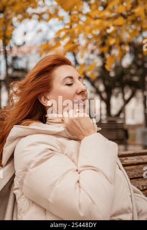 Femme rousse dans la veste gonflante crème se trouve sur le banc du parc, les yeux fermés, profitant de la brise d'automne. Cheveux soufflant dans le vent. Feuilles dorées en arrière-plan. Banque D'Images