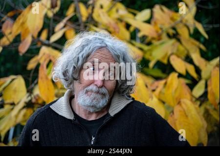 Un homme âgé avec des cheveux gris bouclés et une expression réfléchie se tient devant des feuilles d'automne jaunes vibrantes. Banque D'Images