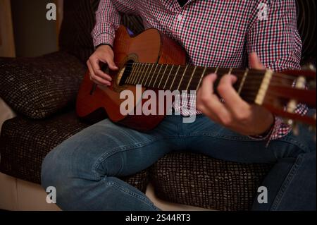 Une vue rapprochée des mains d'une personne caressant une guitare acoustique, portant des vêtements décontractés et assis sur un canapé à motifs, illustrant un e passionné Banque D'Images