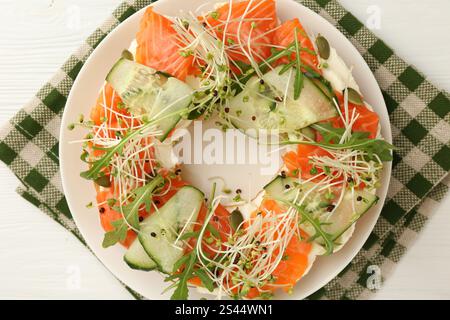 La moitié de délicieux bagel avec saumon, microgreens, roquette et concombre sur une table en bois blanc, vue de dessus Banque D'Images