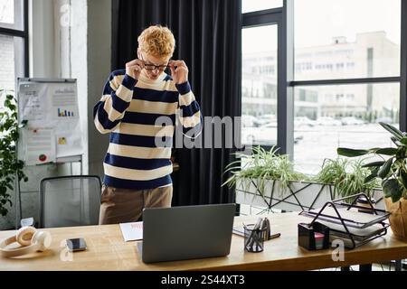 Jeune homme rousse en pull rayé s'engage activement avec son ordinateur portable dans un cadre de bureau élégant. Banque D'Images