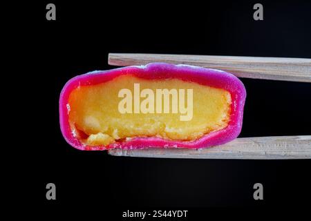 Crème glacée mochi rose avec remplissage de fruits tropicaux jaunes dans des baguettes en bois sur fond noir, gros plan. Le mochi japonais traditionnel est un d inhabituel Banque D'Images
