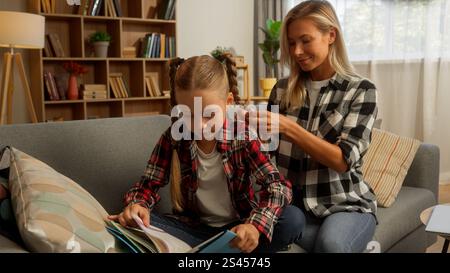 Enfant caucasien petite écolière enfant tenant livre lecture étudier à la maison femme attentionnée mère mère parent tressant cheveux fille lien de famille Banque D'Images