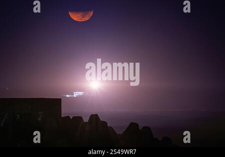 Une lune rouge vif illumine le ciel nocturne au-dessus du phare du cap Fugui. Le faisceau du phare traverse l'obscurité, Taiwan. Côte rocheuse et Banque D'Images