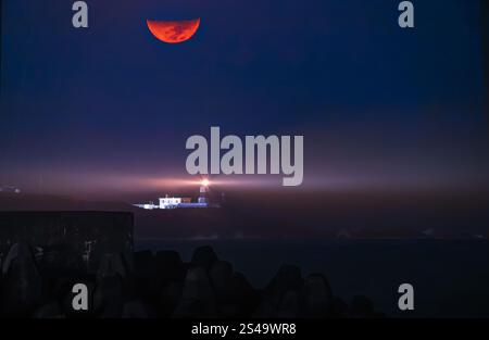 Une lune rouge vif illumine le ciel nocturne au-dessus du phare du cap Fugui. Le faisceau du phare traverse l'obscurité, Taiwan. Côte rocheuse et Banque D'Images
