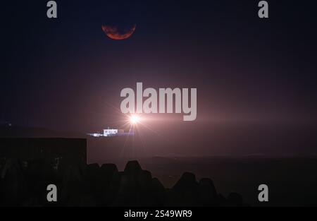 Une lune rouge vif illumine le ciel nocturne au-dessus du phare du cap Fugui. Le faisceau du phare traverse l'obscurité, Taiwan. Côte rocheuse et Banque D'Images