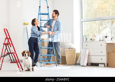 Jeune couple avec escabeau tenant la main et chien Beagle pendant la réparation dans leur nouvelle maison Banque D'Images