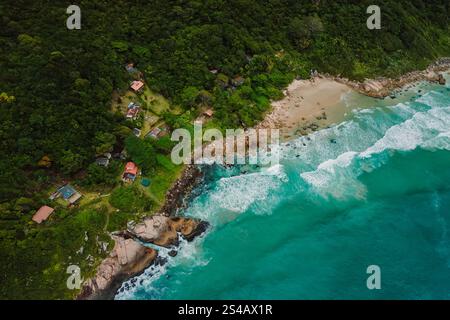 Vue aérienne d'une plage isolée aux eaux turquoises, entourée d'une forêt luxuriante, d'une côte rocheuse et de maisons dispersées au Brésil. Banque D'Images