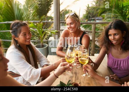 Groupe de femmes diverses toast avec des verres à vin au restaurant. Les amis apprécient le dîner avec des boissons. Réunion sociale, cadre tropical détendu Banque D'Images
