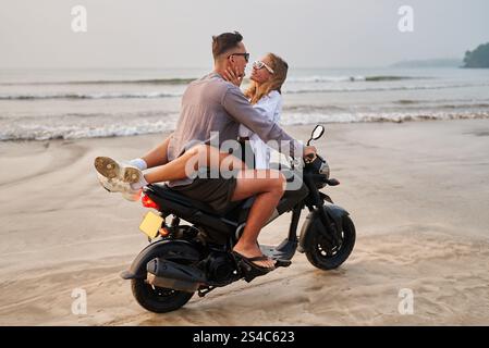 Couple sur moto à la plage. Les amoureux profitent d'un moment romantique sur la rive sablonneuse. Jeune femme embrasse l'homme avec des lunettes de soleil. Vacances, romance, aventure. Datation Banque D'Images