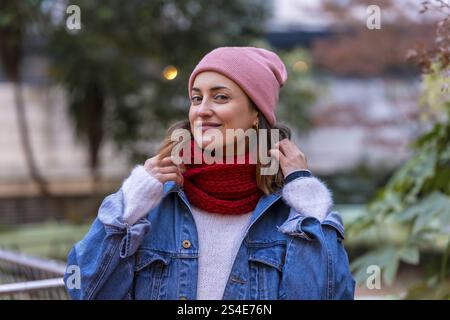 Femme heureuse portant des vêtements d'hiver élégants, ajustant ludique son foulard rouge tout en profitant d'une promenade dans un parc de la ville sereine Banque D'Images