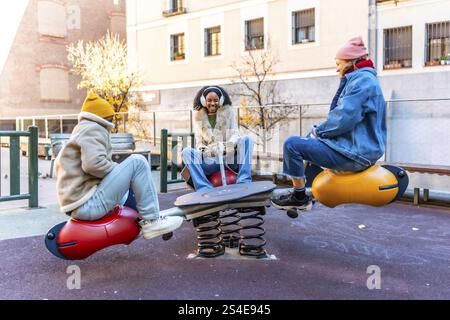 Trois amis profitant d'une journée d'hiver ensoleillée, jouant ensemble sur une balançoire dans une aire de jeux de la ville, s'amusant et riant Banque D'Images