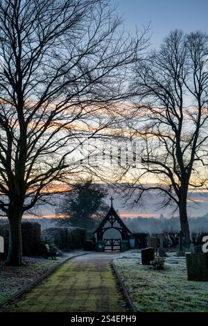 Pierres tombales, porte lych et arbres dans le gel du soir dans un cimetière au crépuscule. Kings Sutton, Northamptonshire, Angleterre. Banque D'Images