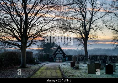 Pierres tombales, porte lych et arbres dans le gel du soir dans un cimetière au crépuscule. Kings Sutton, Northamptonshire, Angleterre. Banque D'Images