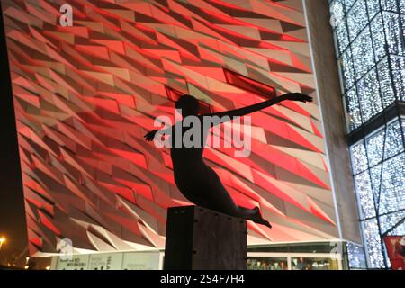 Belfast, Irlande du Nord - 22 décembre 2024 - Silhouette de la sculpture d'art public 'Titanica' à l'extérieur du Titanic Belfast avec des lumières rouges dans le fond. Vue extérieure du bâtiment du musée Titanic Belfast, illuminé la nuit pendant la période des fêtes de Noël dans le quartier Titanic de Belfast. Banque D'Images