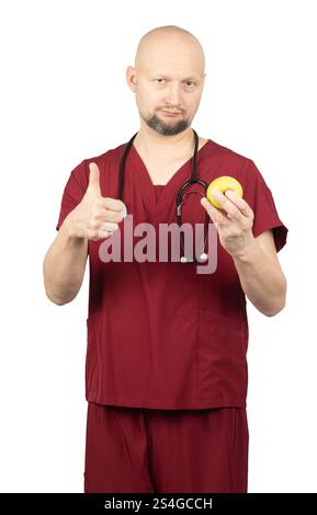 Un médecin chauve avec une barbe, vêtu de gommages rouges et portant un stéthoscope autour du cou, tient une pomme verte dans une main Banque D'Images