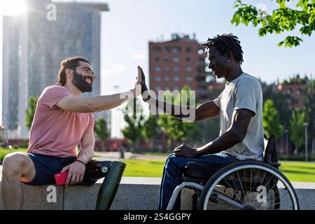 Deux amis, l'un avec une jambe prothétique et l'autre en fauteuil roulant, en haut-fiving dans un parc urbain. L'image exprime l'autonomisation, la diversité et la frite Banque D'Images