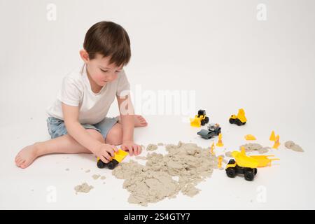 Un enfant souriant joue avec un bulldozer jouet dans le sable cinétique, s'amusant à l'intérieur. Le bébé montre de l'excitation tout en utilisant ses mains pour créer un shap amusant Banque D'Images